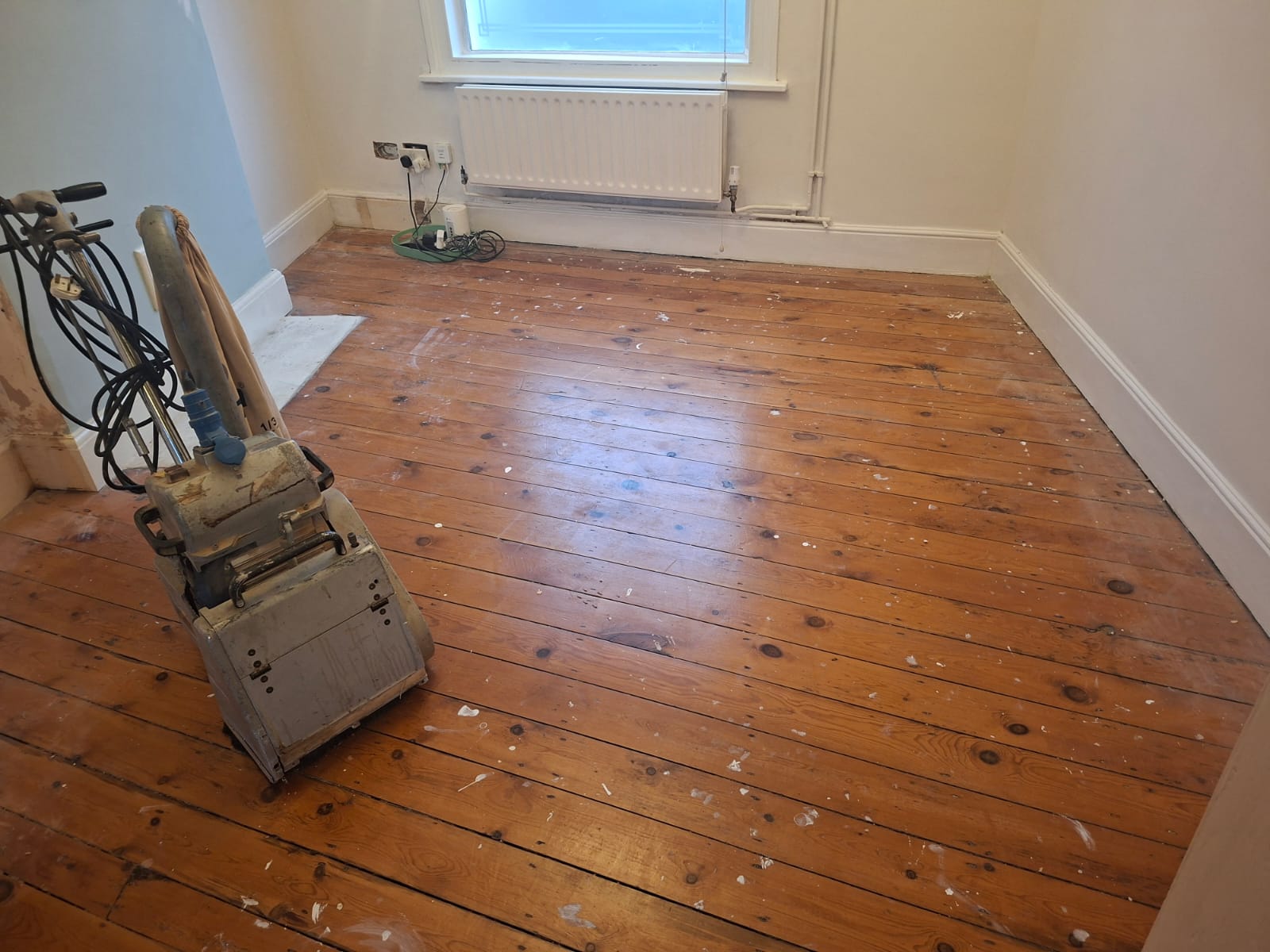 Wood Floor Sanding in Kent on a small bedroom with original pine floorboards, showing a professional sanding machine and paint-splattered boards before restoration.